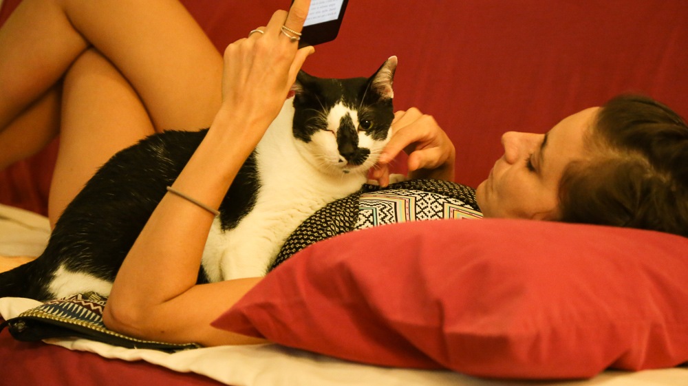 uma mulher deitada em um sofá vermelho, lendo um kindle com um gato preto e branco no colo