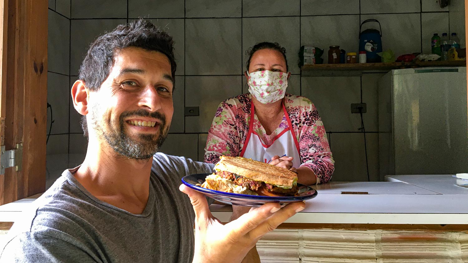 mulher com máscara e homem com um sanduíche na maõ sorrindo na tenda da bike